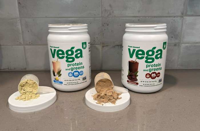 Two open tubs of Vega Protein & Greens protein powder in Chocolate and Vanilla flavors on a kitchen counter